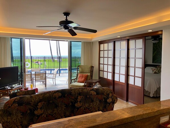 Ein Hotelzimmer mit einer großen Couch, einem Fernseher, einem Deckenventilator, einer Schiebetür und Blick auf einen Pool und Grünflächen.