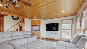 A living room with a sectional sofa, a recliner, a flat-screen TV, wooden ceiling, and a staircase.