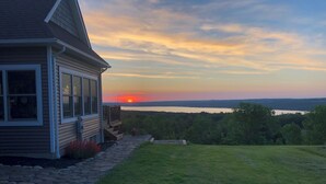 A house with a stone pathway leading to a grassy lawn, overlooking a body of water and a forested area during sunset.