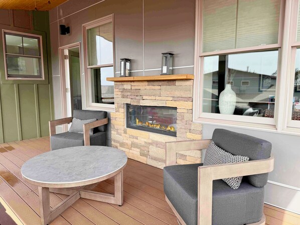 A cozy outdoor seating area with a stone fireplace, wooden furniture, and large windows.