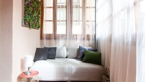 Quarto com cama, mesa de cabeceira, luminária, janela com cortinas leves e planta na parede.
