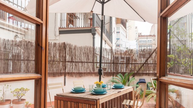 Pátio externo com mesa de madeira posta para refeições, cercado por plantas em vasos e uma cerca de palha.
