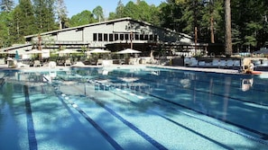Una piscina con agua azul cristalina y separadores de carril, rodeada de sillas de descanso y sombrillas, con un edificio al fondo.