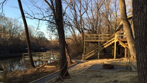 Une terrasse en bois avec un escalier menant à une plateforme surélevée, entourée d’arbres et bordée par un plan d’eau.