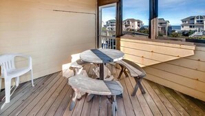 Eine Holzterrasse mit rundem Tisch und Bänken, einem weißen Kunststoffstuhl und großen Fenstern mit Blick auf Gebäude und das Meer.