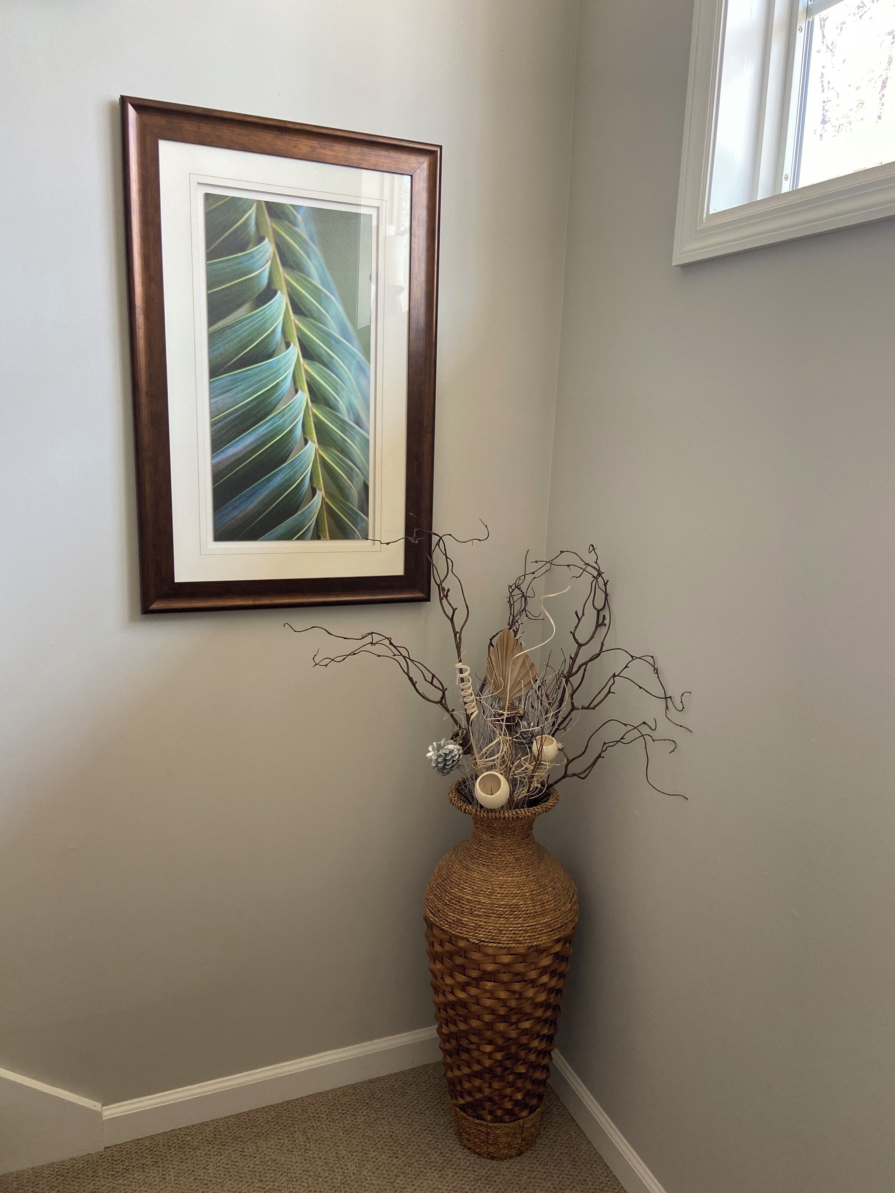 Una impresión botánica enmarcada en una pared, una cesta tejida con ramas decorativas y piñas, y un suelo alfombrado.