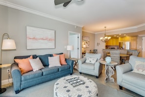 A living room with a blue sofa, a grey armchair, and a round ottoman with a nautical theme. There is a kitchen in the background with yellow cabinets and a dining area with wicker furniture.