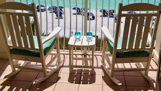 Un balcon avec deux fauteuils à bascule et une petite table sur laquelle sont posés deux verres. La vue donne sur une plage avec des parasols et des gens.