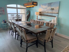 A dining area with a long table, cushioned chairs, a hanging light fixture, and a painting of a landscape.