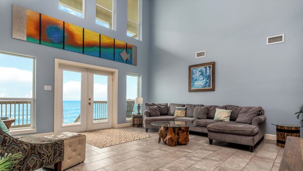 A spacious living room with a large window overlooking the ocean, a sectional sofa, a glass coffee table, and a decorative painting.