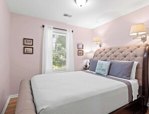 A bedroom with a tufted headboard, a bed with a white quilt, two bedside lamps, a window with white curtains, and framed pictures on the wall.
