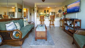 A living room with a sofa, coffee table, and chairs, a kitchen in the background, and a TV on the wall. - Kealia 506 Oceanview Beachfront Resort Great Winter Whale Watching! (Kihei)