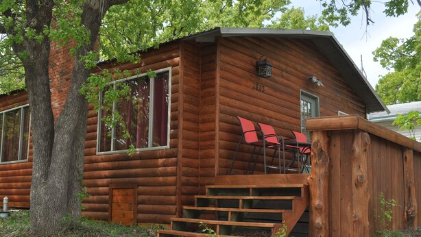 Eine Blockhütte mit Holzfassade, einer Haustür und einer Treppe, die zu einer Veranda mit roten Stühlen führt.