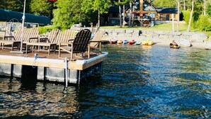 A dock with lounge chairs and a red umbrella, boats, and a house in the background.