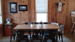 Uma sala de jantar com mesa e cadeiras de madeira, um ventilador e quadros nas paredes.