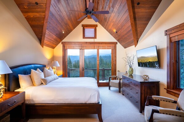 A bedroom with a large bed, wooden ceiling, and a view of mountains through large windows.