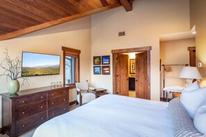 A bedroom with a large bed, wooden dresser, and a desk with a chair. There are framed pictures on the wall and a window with a view of a landscape.
