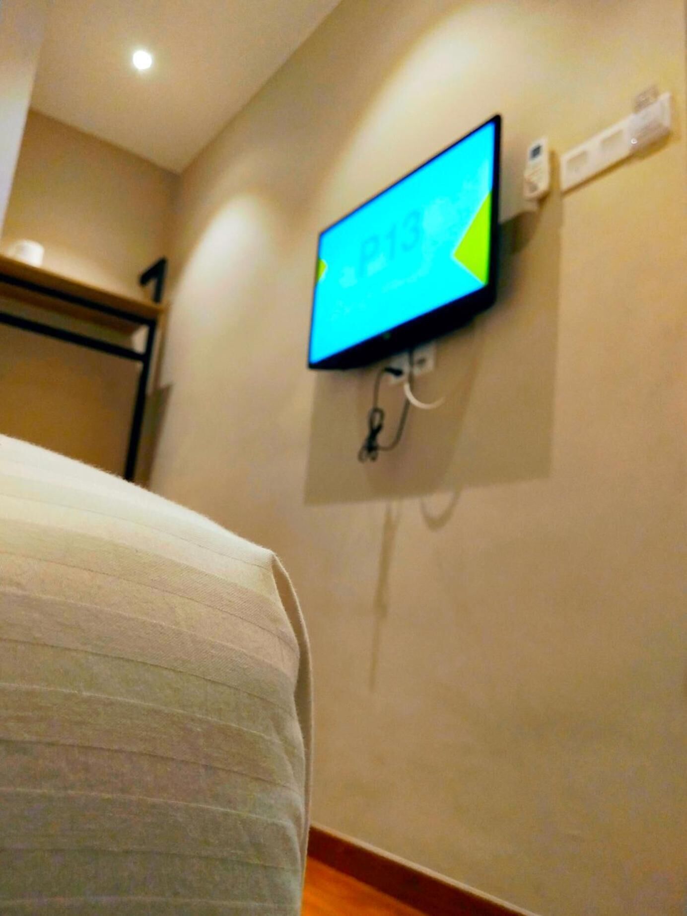 Habitación de hotel con cama, televisor montado en la pared mostrando una pantalla colorida y un enchufe.