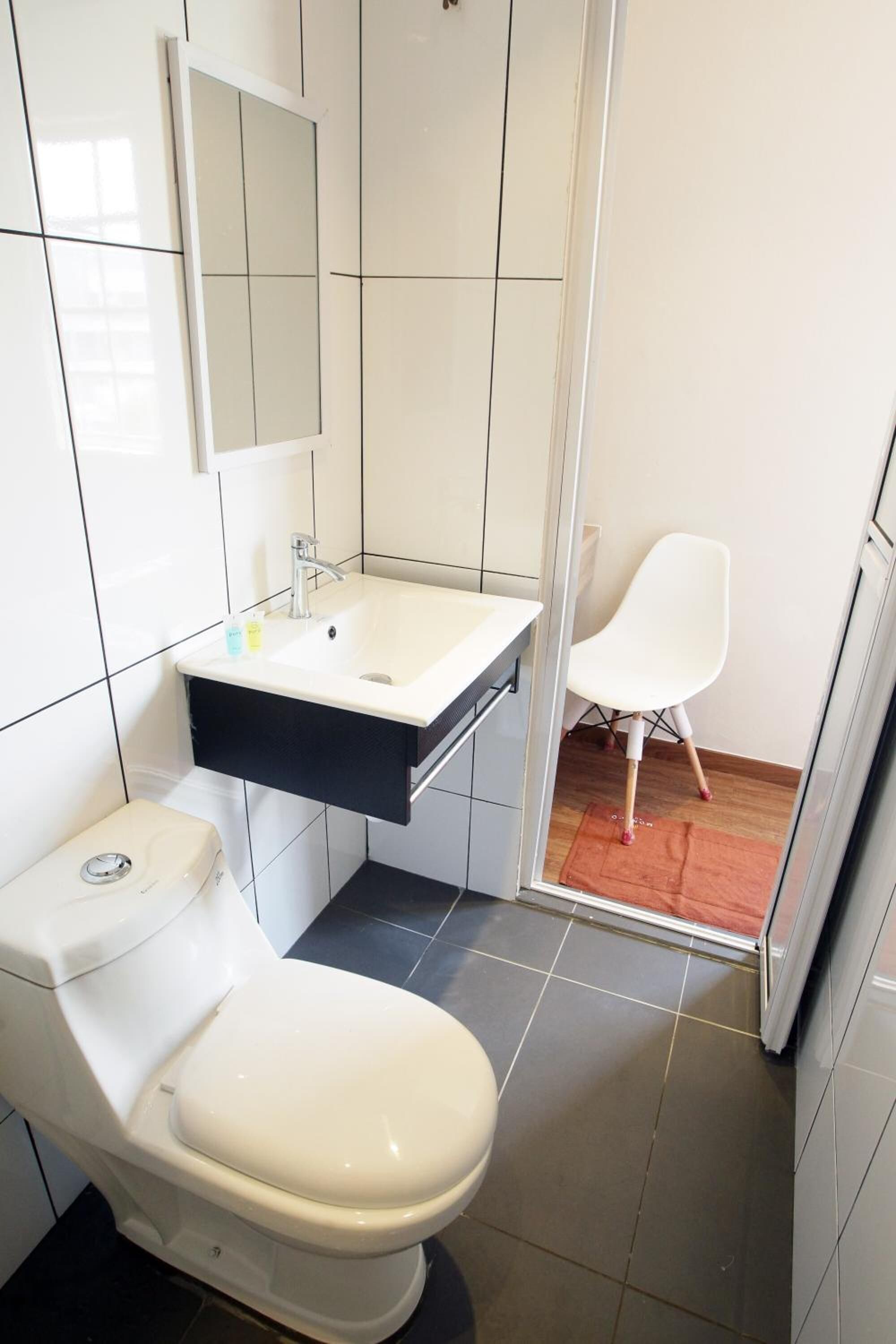 Un baño pequeño con un inodoro blanco, un lavamanos blanco sobre encimera negra y una silla blanca en la esquina.