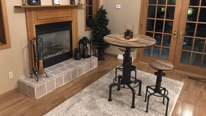 A cozy living room with a fireplace, a round table with two chairs, a clock on the wall, and a door with glass panels.