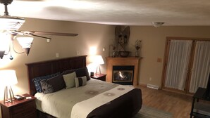 A bedroom with a large bed, a fireplace, a ceiling fan, and a nightstand with a lamp.