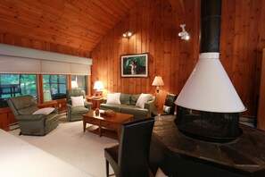 Ein gemütliches Blockhaus-Interieur mit Kamin aus Stein, Holzwänden, einem grünen Sofa, einem Ledersessel und einem Esstisch.