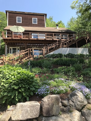Ein zweistöckiges Holzhaus mit einer Terrasse, einer Treppe und einem Garten mit verschiedenen Pflanzen und Blumen.