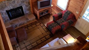 Interior acogedor de una cabaña con chimenea de piedra, un televisor sobre un soporte de madera, un sofá estampado y una escalera de madera.