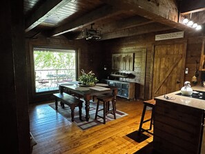 A rustic wooden interior with a dining table, benches, a kitchen area, and a large window providing a view of greenery outside.