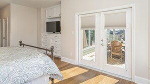 Un dormitorio con una cama, ventilador de techo, televisor y puerta corrediza de vidrio a un balcón con mobiliario exterior.