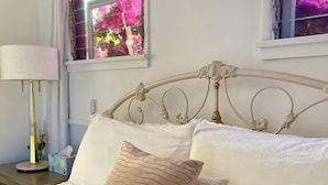 A neatly made bed with white linens and decorative pillows, a bedside lamp, and a window with a view of pink flowers.