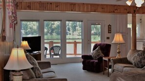 A living room with a television, two lamps, a sofa, a chair, and a view of a deck and greenery outside.