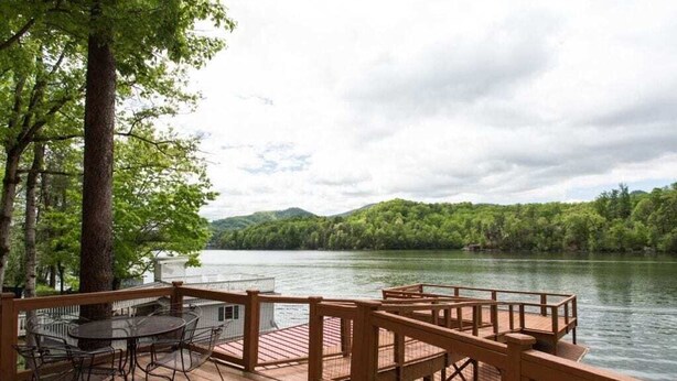 A wooden deck with a table and chairs overlooking a lake, surrounded by trees.