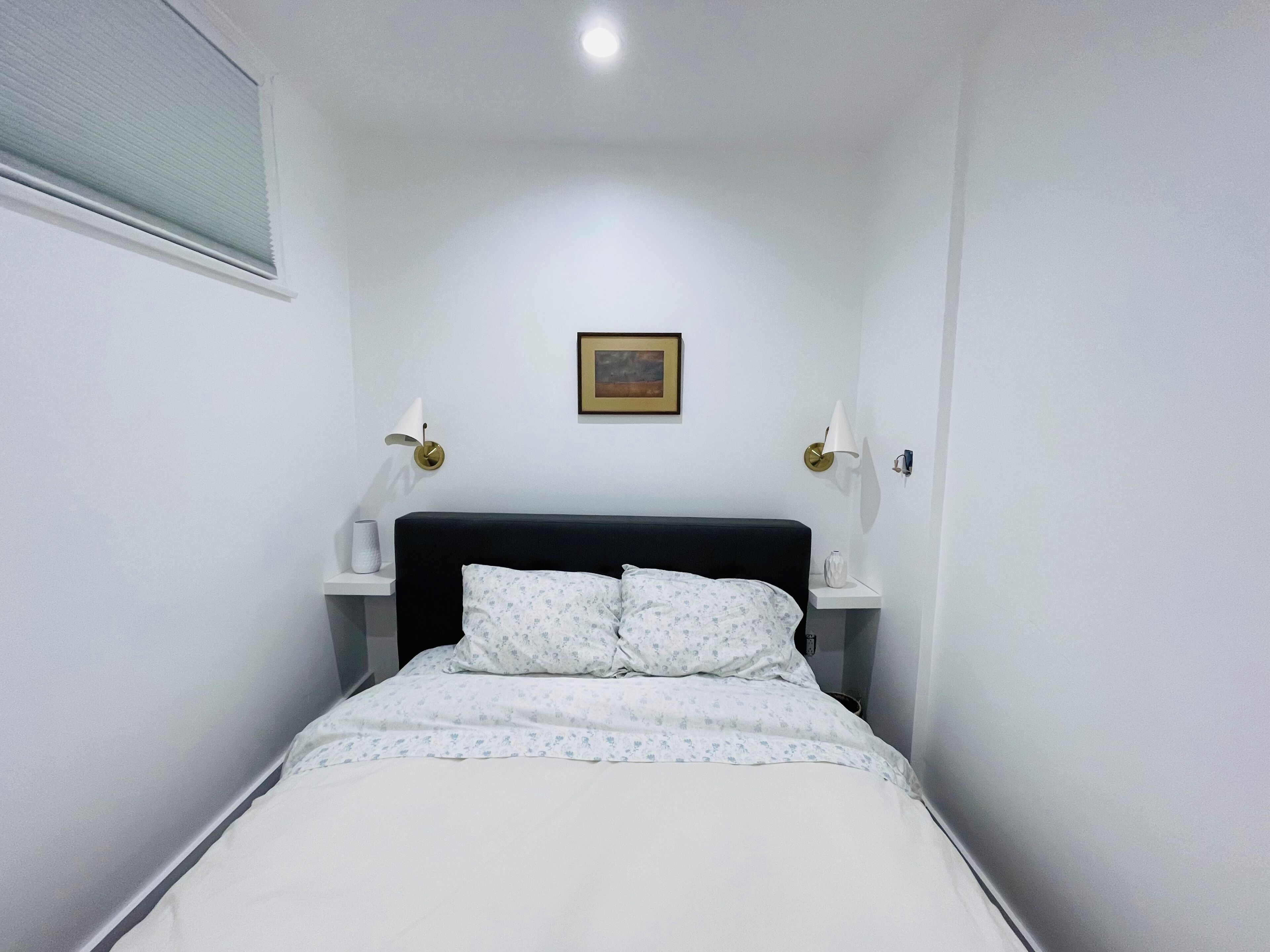 A narrow room with a bed, white walls, a framed picture on the wall, and two bedside lamps.