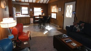 A cozy living room with a red leather chair, a wooden coffee table, and a black leather sofa. There is a dining area with a table and chairs, and a kitchen in the background.