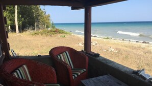 A covered seating area with wicker chairs and a wooden table, overlooking a beach with waves and a clear sky. - Rustic Authentic Michigan Log Cabin (Beaver Island)