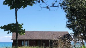 A cabin with a stone foundation and a wooden exterior, surrounded by tall grass and trees, overlooking a body of water. - Rustic Authentic Michigan Log Cabin (Beaver Island)