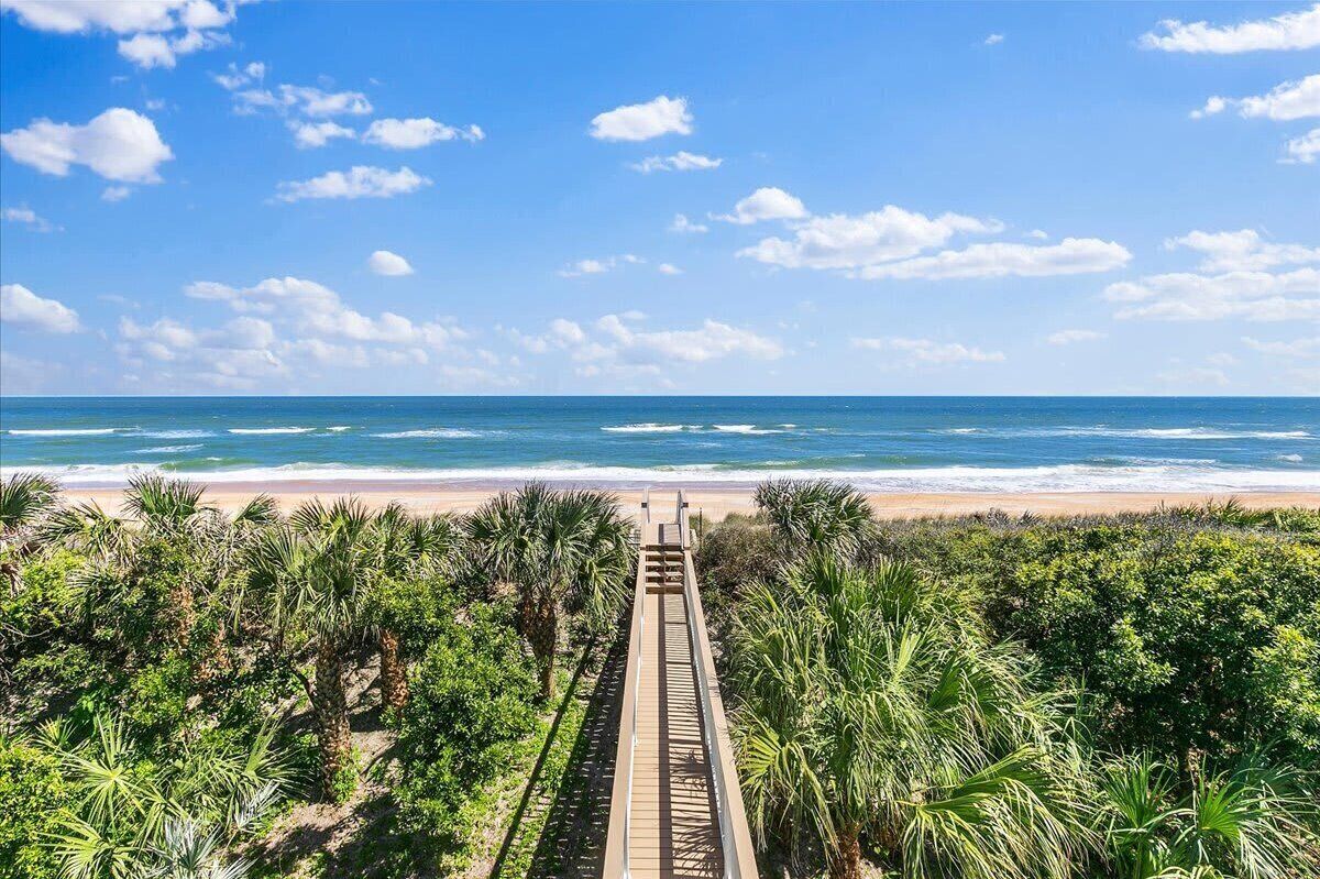 A beach with a wooden boardwalk leading to the ocean, surrounded by dense greenery.