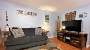 Un soggiorno con un divano grigio, cuscini decorativi, un televisore a schermo piatto su un mobile in legno, un tappeto ornamentale e opere d'arte alle pareti, tra cui un cartello con la scritta "THE LAKE HOUSE".