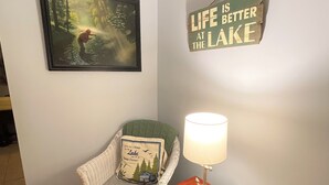 Una stanza con un quadro raffigurante una scena di bosco, una sedia con un cuscino, un cartello con la scritta 'LIFE IS BETTER AT THE LAKE' e un tavolo con una lampada.