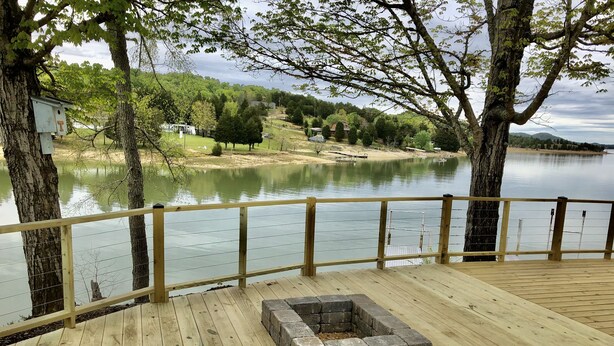Un'area prendisole in legno con un braciere, affacciata su un lago tranquillo con alberi e case sullo sfondo.