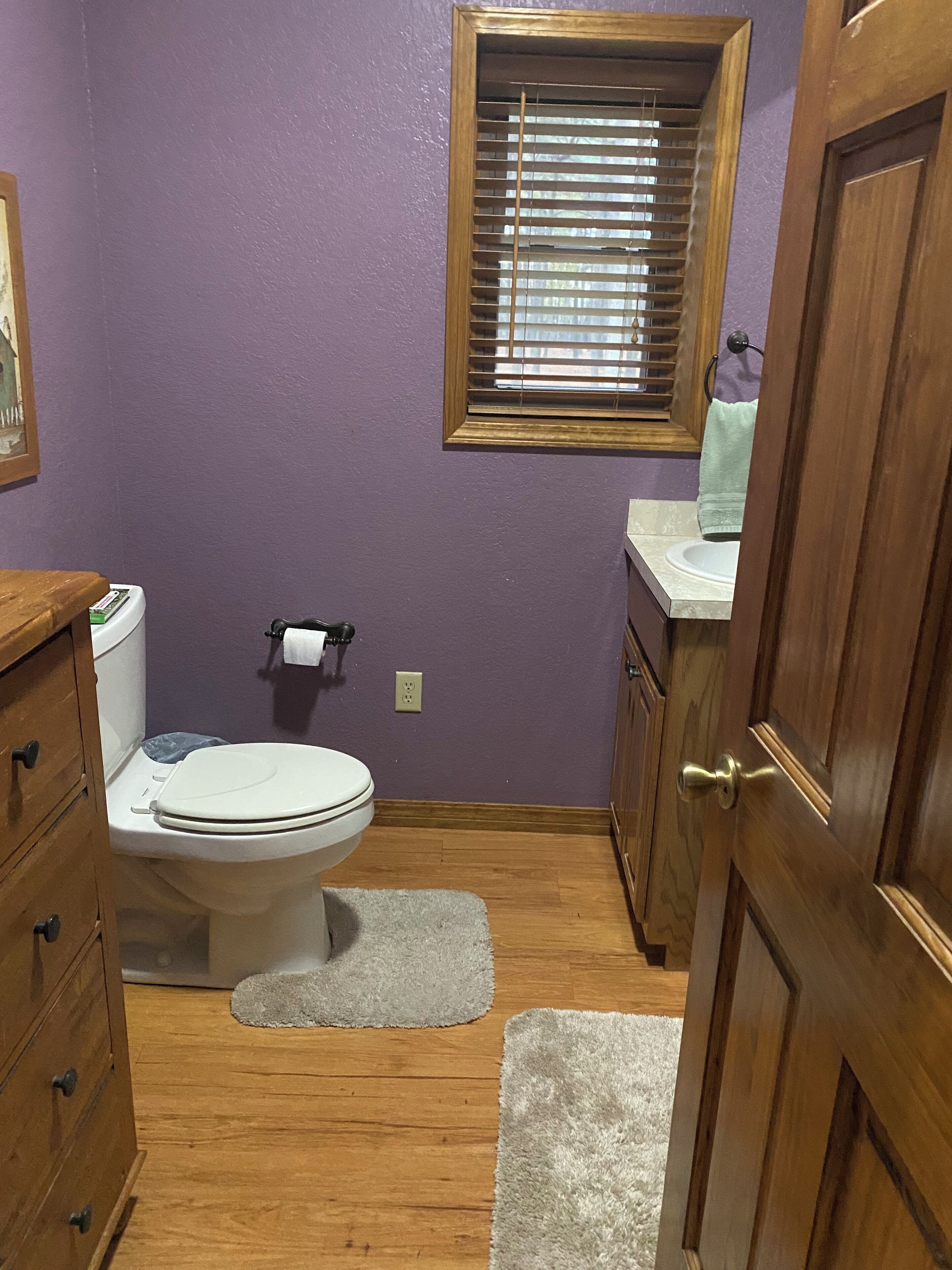 Une salle de bain avec des toilettes, des meubles en bois, une fenêtre avec des stores et une porte.