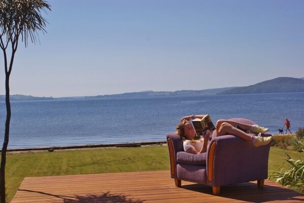 A person relaxing on a purple chair by a wooden deck overlooking a body of water with distant hills. - Lounge to Lake - Water's Edge, Lake Rotorua (Ngongotaha)