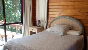 A bed with a woven headboard, a bedside table with a lamp, and a view of the outdoors through a large window.