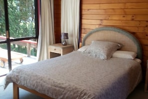 Uma cama com cabeceira em tecido, uma mesa de cabeceira com abajur e vista para o exterior através de uma janela grande.