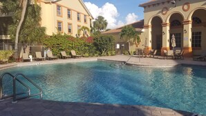 A swimming pool with lounge chairs and umbrellas, surrounded by palm trees and a building with arches. - Beautiful Naples Condo (Naples)