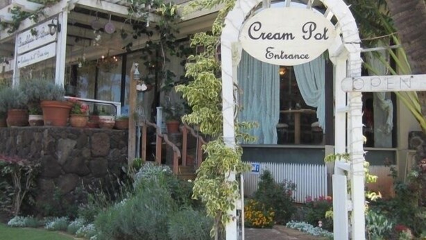 Entrada do Cream Pot com uma placa, rodeada por vasos de plantas e um muro de pedra.