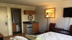 Quarto com cama, almofadas, mesa de cabeceira, escrivaninha, cadeira, televisão, cozinha e banheiro.