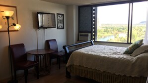 Quarto com cama, escrivaninha, duas cadeiras, televisão e janela com vista para um lago e árvores.