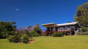 Une maison à deux étages avec une terrasse, entourée de verdure et d’arbres.